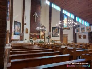 Wnętrze kościoła. Widok na rzędy ławek kościelnych ustawionych w regularnych szeregach. W tle widoczny jest ołtarz.