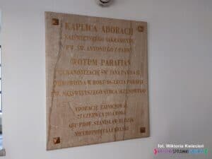 Tablica informacyjna z piaskowego granitu zamocowana na białej ścianie. Zawiera inskrypcję: „Kaplica Adoracji Najświętszego Sakramentu pod wezwaniem św. Antoniego z Padwy. Wotum parafian za kanonizację św. Jana Pawła II, odnowiona w roku 80-lecia parafii pw. Najświętszego Serca Jezusowego. Adoracje zainicjował 27 czerwca 2014 r. abp prof. Stanisław Budzik, Metropolita Lubelski.”