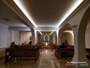 Wnętrze Kaplicy Adoracji w przyjemnym półmroku, oświetlone ciepłym światłem przysufitowym. Ujęcie z perspektywą na ołtarz; po obu stronach rzędy ławek, w których w różnych odległościach siedzą trzy osoby