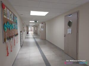 Długi, jasnooświetlony korytarz szkolny. Po prawej stronie ciąg drewnianych drzwi prowadzących do sal lekcyjnych, po lewej stronie na ścianach wiszą tablice informacyjne