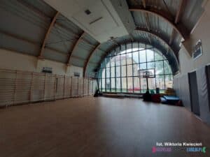 Przestronne wnętrze sali gimnastycznej z łukowatym sufitem i drewnianą podłogą. Na ścianach zamontowane są rzędy drewnianych drabinek gimnastycznych