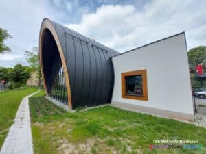 Zewnętrzna elewacja frontowa sali gimnastycznej z wysokim, półokrągłym dachem i przeszkloną przednią ścianą budynku