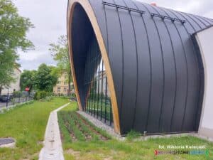 Zbliżenie na zewnętrzną elewację frontową sali gimnastycznej z wysokim, półokrągłym dachem i przeszkloną przednią ścianą budynku