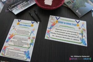 Na ciemnobrązowym stole leżą dwa plakaty. Pierwszy - informuje o wystawie prac plastycznych uczniów ze Szkoły Specjalnej nr 26 im. Janusza Korczaka w Lublinie. Drugi plakat przedstawia instrukcje głosowania na najciekawszą pracę, zachęcając do oddania swojego głosu. Plakaty są kolorowe, a ich tekst jest czytelny