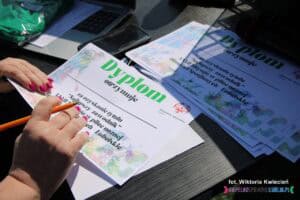 Zbliżenie na kobiece dłonie z różowymi paznokciami, które trzymają brzeg dyplomu otrzymywanego za uzyskanie tytułu 'Najlepszy zawodnik' XXXII Mityngu w piłce nożnej. W prawej ręce znajduje się długopis, gotowy do złożenia podpisu