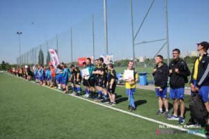 Prezentacja drużyn i zawodników. Na słonecznym boisku przed białą linią boczną ustawili się zawodnicy w różnobarwnych strojach sportowych