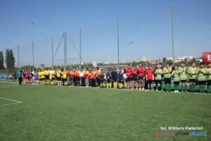 Prezentacja drużyn i zawodników. Na słonecznym boisku w długim szeregu stoją zawodnicy w różnobarwnych strojach sportowych
