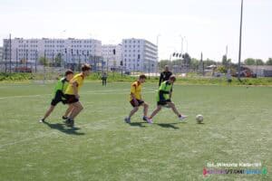 Na słonecznym boisku piłki nożnej trwa dynamiczna akcja pomiędzy czterema zawodnikami. Gracze są ubrani w kolorowe koszulki i krótkie spodenki. Jeden z nich znajduje się przy piłce, pozostali za nim. Boisko jest zielone, w tle widać sędziego, bramkę oraz bloki mieszkalne.