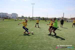 Na boisku piłki nożnej trwa dynamiczna akcja pomiędzy czterema zawodnikami. Jeden z nich znajduje się przy piłce, dwóch innych, reprezentujących przeciwne drużyny, stara się ją przejąć, a obrońca jest gotowy do interwencji. Jeden zawodnik jest ustawiony nieco dalej, obserwując całą sytuację. Boisko jest zielone, w tle widać bramki oraz kilku kibiców. Słońce oświetla boisko, a gracze są ubrani w kolorowe koszulki i krótkie spodenki