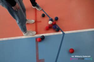 Na podłodze hali widoczny jest układ czerwonych i niebieskich buli, rozmieszczonych wokół białego jacka. Obok dwie pary nóg w obuwiu sportowym