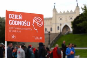 Na pierwszym planie symbol Dnia Godności - pomarańczowa chorągiewka z nazwą uroczystości. W dalszej części kadru zgromadzeni ludzie na tle zamku