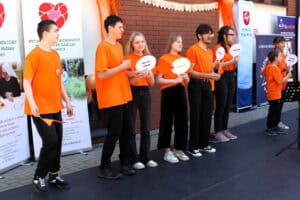 Na scenie ośmioosobowa grupa młodzieży w pomarańczowych koszulkach prezentująca utwór muzyczny. Niektóre osoby trzymają w rękach tabliczki z napisami, np. „Nie lubię swetrów”