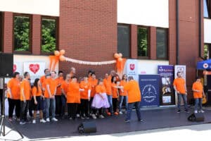 Na scenie młodzi artyści prezentujący utwór muzyczno – taneczny. Przed grupą artystów śpiewających para wykonuje taniec. Wszyscy mają na sobie pomarańczowe koszulki