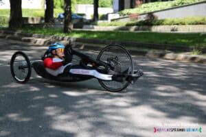 Zawodnik na handbike’u w kasku i stroju sportowym. Wzdłuż prawej nogi widoczny czerwony napis 'Polska' na białym tle.