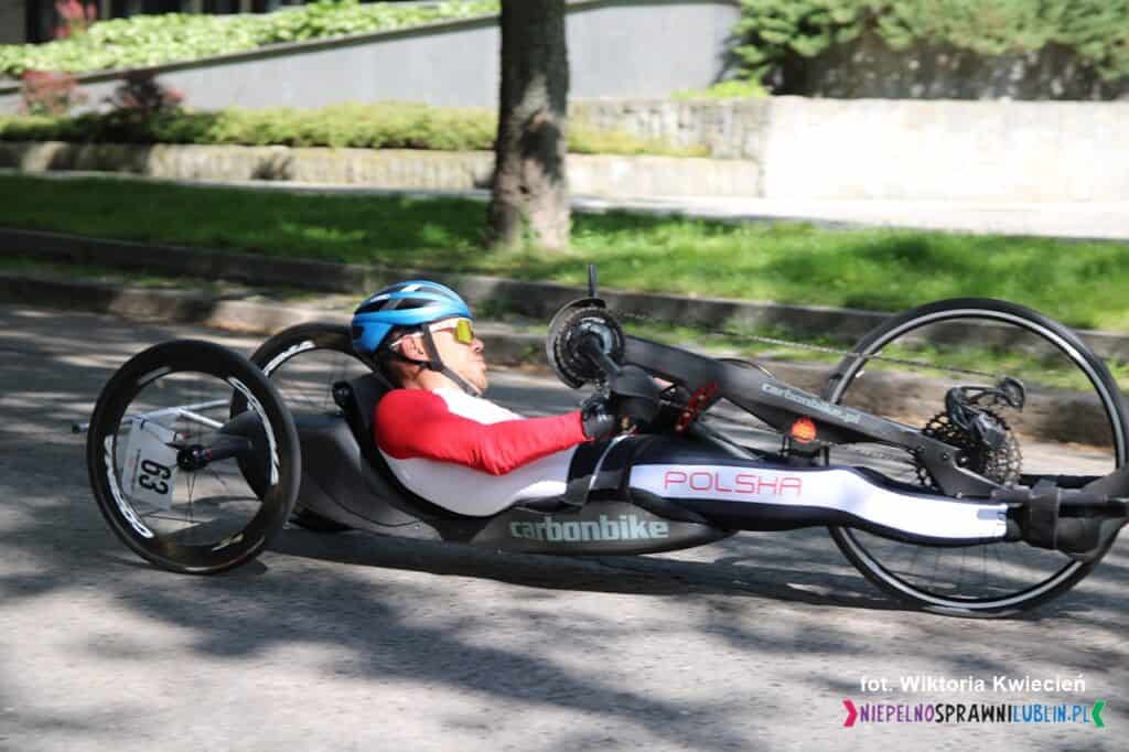 Zawodnik z numerem 63 poruszający się na handbike’u w trakcie jazdy, bliskie ujęcie z boku. Ma na sobie kask i sportowy strój, ręce na korbach napędzających rower