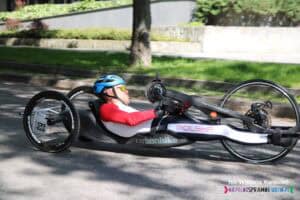 Zawodnik z numerem 63 poruszający się na handbike’u w trakcie jazdy, bliskie ujęcie z boku. Ma na sobie kask i sportowy strój, ręce na korbach napędzających rower