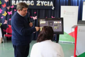 Od lewej strony kadru: Piotr Celmer-Domański (AssisTech) prezentujący możliwości C-Eye. Przed urządzeniem siedzi jedna ze słuchaczek, która zapoznaje się z możliwościami urządzenia. Na monitorze urządzenia widok na stronę główną menu