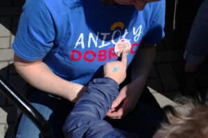 Zbliżenie na dłoń dziecka wskazującą palcem na przypinkę Dnia Godności, która jest na tle niebieskiej koszulki wolontariuszki Anioły Dobroci. Przypinka ma napis „Dzień Godności Osób z Niepełnosprawnością Intelektualną”