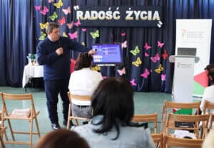 Widok ogólny na prezentację technologii IT. Od lewej: Piotr Celmer-Domański (AssisTech) prezentujący przed jedną ze słuchaczek (siedzącą przed urządzeniem) jego możliwości. Po prawej, na tle granatowej kurtyny baner informacyjny PFRON