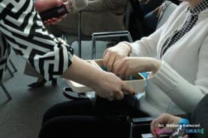 Zbliżenie na prawą rękę trzymającą drewniane pudełko z losami; w jego wnętrzu inna para rąk przeszukuje zawartość.