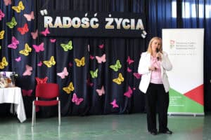 W prawej części kadru Agnieszka Słoboda - kierownik OWiT Łęczna, podczas wystąpienia. W tle baner informacyjny PFRON oraz granatowa kurtyna z motylami w różnych kolorach (żółte, zielone, różowe) i napisem „Radość życia”