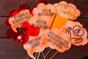 Pomarańczowe gadżety do zdjęć: okulary w kształcie serc i chorągiewki z hasłami przewodnimi uroczystości, np.: „Jestem wyjątkowy”, „Dzień Godności Osób z Niepełnosprawnością Intelektualną”, „Jestem wartościowy”