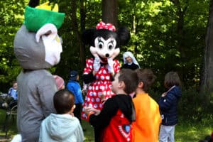 Zabawa dzieci w lesie, z osobami przebranymi za postacie z bajek. W centralnej części kadru Myszka Minni i otaczające ją dzieci. Obok siostra zakonna