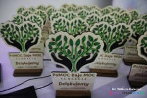 Statuetka wykonana w drewnie. Zawiera kolorowe logo Fundacji PoMOC Daje MOC, napis "Dziękujemy" oraz datę "17-18.05.2025"