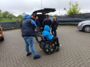 Pięć osób na parkingu, wśród nich jedna na wózku inwalidzkim, stoją przed otwartym bagażnikiem samochodu typu VAN