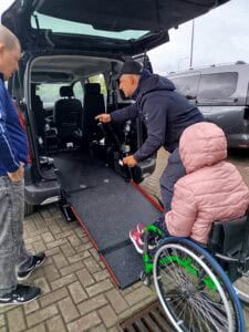 Osoba na wózku inwalidzkim wjeżdża po rozłożonej platformie do wnętrza samochodu w asyście dwóch mężczyzn