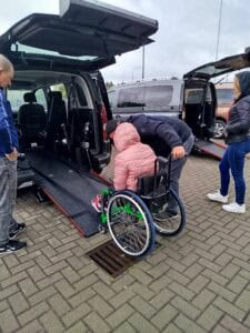 Osoba na wózku inwalidzkim wjeżdża do wnętrza samochodu po rozłożonej platformie. Towarzyszy jej dwóch mężczyzn udzielających pomocy, z boku przygląda się kobieta