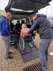 Jedna osoba na wózku inwalidzkim wjeżdża po rozłożonej platformie do wnętrza samochodu. Wózek pcha jeden z mężczyzn, drugi stoi obok i przygląda się z uwagą, gotów do pomocy