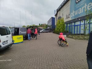 Ogólny widok na plac przed Centrum Wózków Inwalidzkich, gdzie prezentowano samochody dostosowane do potrzeb osób na wózkach. Grupy osób oglądają pojazdy