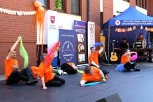 Na scenie siedmioosobowa grupa młodzieży podczas występu. Część artystów leży, pozostałych czworo siedzi i macha kolorowymi chustami