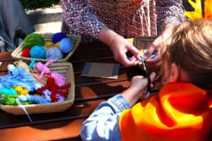 Na zdjęciu warsztat rękodzielniczy. Zbliżenie na dłonie dziecka i opiekuna podczas przygotowywania kukiełki z włóczki. W lewej części kadru dwa koszyki: z kolorową włóczką i gotowymi kukiełkami