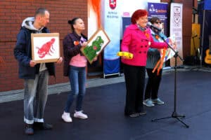 Czteroosobowa grupa dorosłych artystów podczas występu muzycznego. Na pierwszym planie solistka z żółtym tamburynem. Pozostałe osoby tuż za nią, trzymają w rękach kolorowe rysunki zwierząt: raka, żaby i ryby