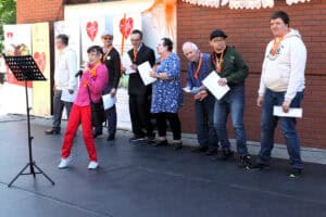Na scenie ośmioosobowa grupa podczas wykonywania utworu muzycznego. Na pierwszym planie solistka z mikrofonem. Wszyscy noszą pomarańczowe zawieszki w kształcie serc lub kwiatów