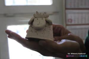 Zbliżenie na fantazyjnego, wielkanocnego ceramicznego baranka z ulepioną sterczącą grzywką, stojącego na rozłożonej damskiej dłoni