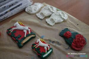 Gipsowe odlewy wykonane w pracowni rękodzieła: pomalowane koziołki z herbu Lublina oraz cztery elementy z motywem kwiatów — jeden pomalowany i trzy surowe, białe