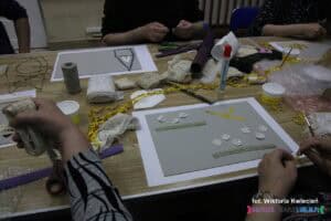 Zbliżenie na stół, na którym panuje artystyczny rozgardiasz — porozrzucane są różnorodne materiały strukturalne, koronki, tasiemki i sznureczki. Widać ręce uczestników warsztatów rękodzielniczych, którzy pracują nad tworzeniem matryc