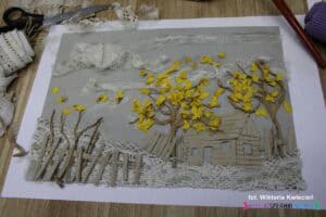 Matryca do kolografii przedstawiająca domek i płotek z tektury falistej oraz drzewa z papierowego sznurka z tekstylnymi liśćmi z ozdobnej tasiemki, nad krajobrazem unoszą się koronkowe chmury