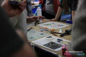 Wokół stołu stoją uczestnicy. Na stole leży matryca częściowo pokryta farbą, nad którą unosi się ręka z tubką farby drukarskiej. Pośrodku stołu rozłożone są różne przybory, a w tle widać kolejną matrycę