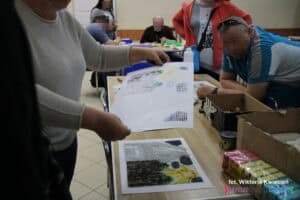 Wokół stołu stoją uczestnicy. Częściowo widoczna prowadząca trzyma w dłoni papier z odbitką wykonaną od leżącej na stole matrycy