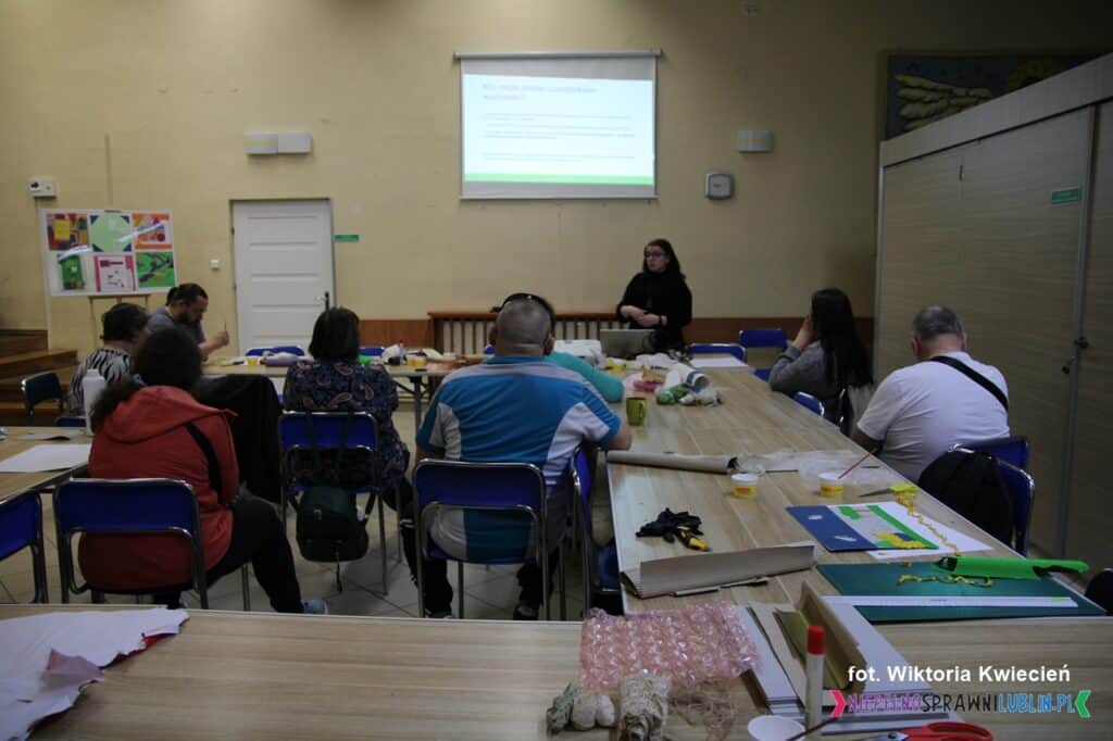 Na pierwszym planie widoczny jest stół z artystycznym rozgardiaszem po zakończonych warsztatach rękodzielniczych. W tle pani Katarzyna prowadzi dla grupy uczestników, widzianych od tyłu, prezentację multimedialną wyświetlaną na ekranie przymocowanym do ściany
