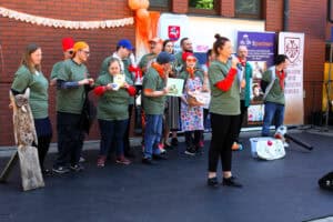 Widok na scenę podczas występu młodych artystów. Wszyscy są ubrani w koszulki w odcieniu leśnej zieleni. Niektórzy mają na głowach kolorowe czapki – kaszkietówki