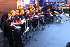 Na scenie młodzież śpiewająca i grająca na instrumentach muzycznych pod okiem siostry zakonnej. Młodzi artyści mają na sobie granatowe koszulki, pomarańczowe chusty i pomarańczowe „kaszkietówki”, odwrócone daszkiem do tyłu