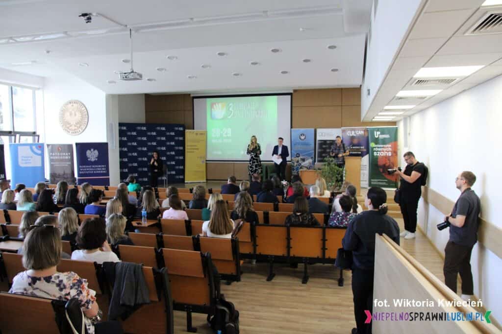 Widok ogólny na salę konferencyjną KUL z perspektywy widowni. Na scenie znajduje się prowadzący konferencję oraz tłumacz PJM. W tle stoją rollupy reklamowe współorganizatorów i partnerów