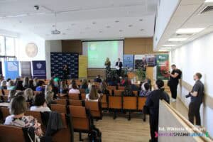 Widok ogólny na salę konferencyjną KUL z perspektywy widowni. Na scenie znajduje się prowadzący konferencję oraz tłumacz PJM. W tle stoją rollupy reklamowe współorganizatorów i partnerów