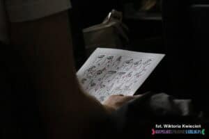 Zbliżenie na rękę uczestnika warsztatów trzymającą wydruk alfabetu manualnego (palcowego) – zbiór znaków migowych (daktylogramów) odpowiadających literom alfabetu