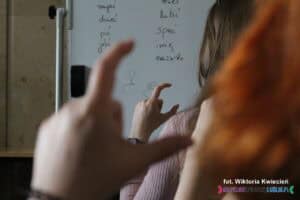 Uczestnicy powtarzają palcami znak litery „C” w języku migowym, pokazany przez dr Aleksandrę Borowicz. Uczestnik na pierwszym planie jest rozmazany, a ostrość zdjęcia skupiona na dłoni osoby siedzącej dalej. W tle widoczna jest tablica z wyrazami zaplanowanymi do nauki podczas warsztatów